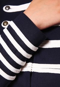 Granatowy mankiet kurtki z białym, splecionym wykończeniem i ozdobnymi guzikami. Charakteryzuje się fakturowaną tkaniną z wyraźnymi poziomymi paskami.