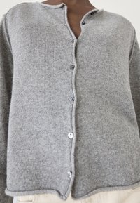 Cardigan grigio lavorato a maglia con scollatura a V, con chiusura a bottoni e texture a coste. Dettagli includono bottoni tondi e orlo leggermente curvo.