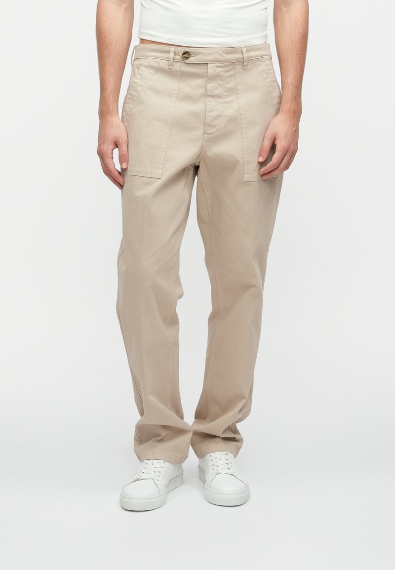 Beige auduma bikses ar brīvu piegriezumu, ar divām priekšējām kabatām, pogu aizdari un smalkām šuvju detaļām. Kopā ar baltiem sporta apaviem.