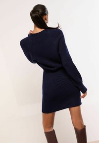 Sötétkék, kötött pulóverruha bordázott textúrával, hosszú ujjakkal és kényelmes szabással. Az alsó szegély a térd fölött ér véget.