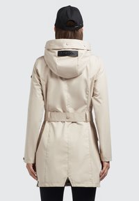Beige vedenkestävä takki, jossa on huppu ja vyötäröllä solki. Takissa on sileä pinta, napit ja logo ommeltuna taakse.
