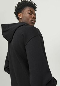 Black hoodie with a drawstring, relaxed fit, and textured fabric. Features a hood and dropped shoulder seams, viewed from the back.