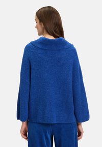 Niebieski sweter o teksturowanym splocie z szerokim kołnierzem i dzwonowymi rękawami, charakteryzujący się luźnym krojem i żebrowanym wzorem. Noszony z dopasowanymi niebieskimi spodniami.