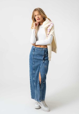 Fustă maxi din denim cu slit frontal, detalii cu nasturi și culoare bleu deschis. Asortată cu un top alb cu mânecă lungă și un pulover crem.
