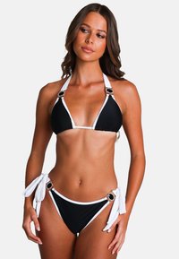 Frau, die einen schwarz-weißen Halterneck-Bikini mit dekorativen Schnallen trägt, steht vor einem schlichten weißen Hintergrund.