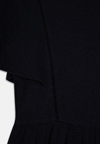 Černá texturovaná tkanina se strukturovaným designem, s volánky na spodním lemu a viditelným švem po straně.