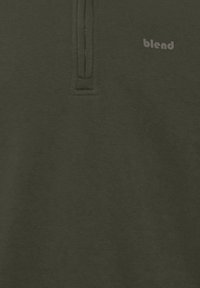 Suéter verde caqui con media cremallera y cuello alzado; cuenta con un sutil logo de la marca en gris claro en la parte izquierda del pecho. Textura suave.