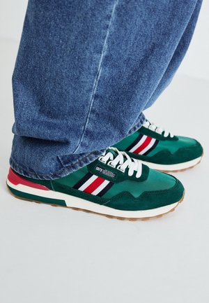 Baskets vertes avec lacets blancs et rayures rouges, blanches et noires, portées avec un jean en denim bleu sur un sol blanc.