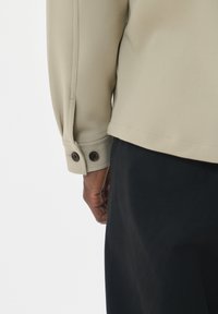 Camicia beige a maniche lunghe con polsini abbottonati, texture del tessuto liscia e orlo dritto, abbinata a pantaloni scuri.