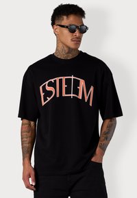 T-shirt noir oversize avec un graphique 'ESTEEM' en rouge. Fait en coton, avec des manches courtes et un col rond. Texture lisse.