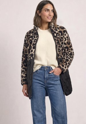 Femme portant une veste imprimé léopard avec des panneaux matelassés noirs, un pull blanc côtelé et un jean bleu, debout avec une main dans la poche.