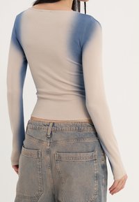 Maglietta a maniche lunghe beige con effetto ombre blu, caratterizzata da un design aderente e scollatura arrotondata. Abbinata a jeans di denim blu chiaro.