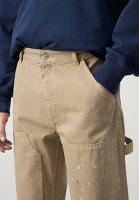 Persona que lleva pantalones de trabajo beige salpicados de pintura con bolsillos laterales y una camiseta de manga larga azul oscuro, una mano en el bolsillo y la otra relajada.