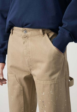 Persona que lleva pantalones de trabajo beige salpicados de pintura con bolsillos laterales y una camiseta de manga larga azul oscuro, una mano en el bolsillo y la otra relajada.