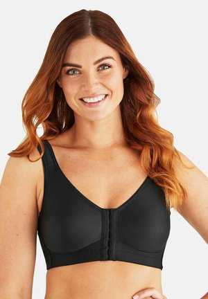 Lachende vrouw met golvend rood haar die een zwarte sportbeha met voorhaak draagt, staand tegen een effen lichte achtergrond.
