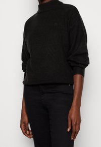 Pull en maille noir avec un col montant et des manches larges, présentant une texture subtile et un petit logo sur la poitrine. Associé à un pantalon noir.