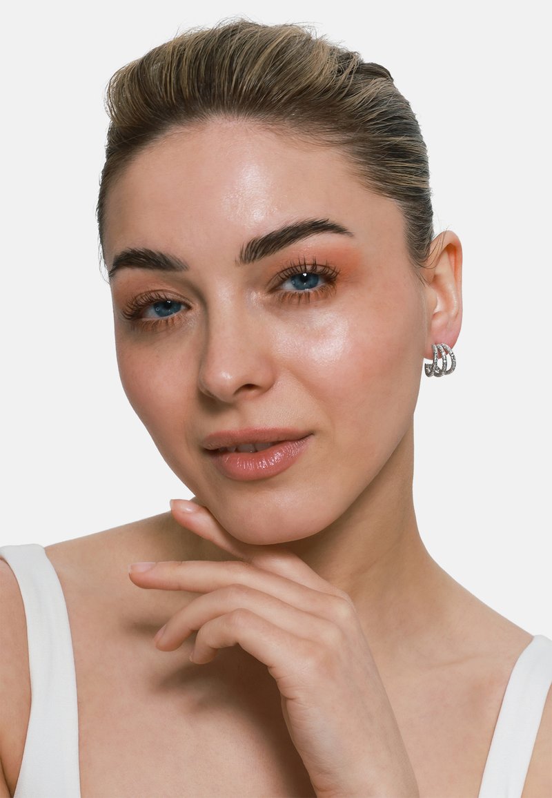 Silberne Creolen mit aufwendigem verdrehtem Design, präsentiert an einer Person mit klarer Haut und leichter Make-up. Weißes Oberteil und neutraler Hintergrund.