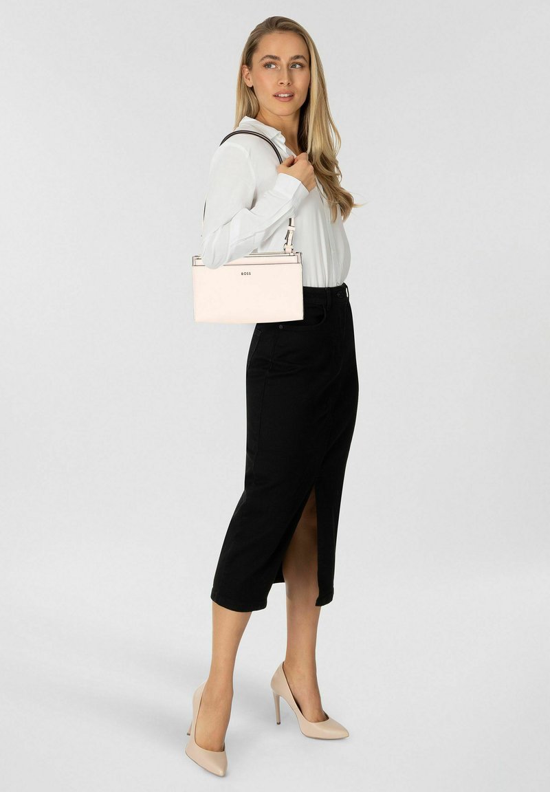 Femme en blouse blanche, jupe midi noire avec fente avant, talons nus, tenant un sac à main beige clair sur l'épaule, debout devant un fond uni.