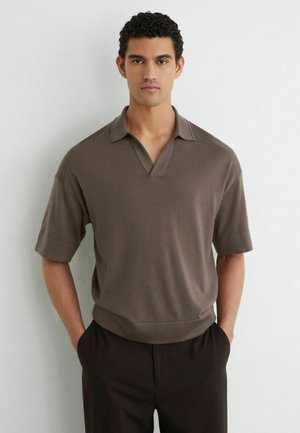 Uomo con capelli corti e ricci che indossa una camicia marrone a maniche corte con colletto e pantaloni scuri, in piedi contro un muro bianco semplice.