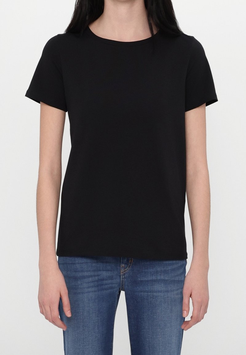 Femme portant un t-shirt noir uni à manches courtes et un jean bleu, debout devant un fond blanc uni.