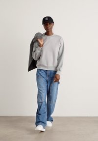 Felpa grigia con polsini a costine, jeans blu larghi e sneakers bianche. Indossa un cappellino nero e tiene in mano una giacca grigia. Sfondo neutro.