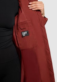 Persona vestida con un conjunto negro sostiene un abrigo largo acolchado en color óxido, mostrando un bolsillo interior con la etiqueta en blanco y negro "Khujo Tim original para unos pocos".