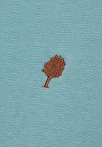 Tessuto turchese con una superficie liscia presenta un piccolo design di un albero ricamato in marrone. Dettaglio semplice e minimalista.