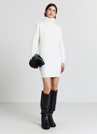 Vestito maglione bianco a coste con collo alto e maniche lunghe, abbinato a stivali neri al ginocchio e pochette nera. Tessuto liscio.