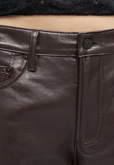 Podrobni posnetek temno rjavih usnjenih hlač, pritrjenih s črnim gumbom, nošenih s črnim čipkastim topom, ki je viden nad pasom.