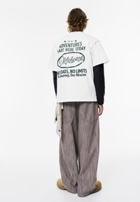 Hvid kortærmet t-shirt med grønne trykte bogstaver på ryggen, parret med stribede, vide grå bukser og beige sko.