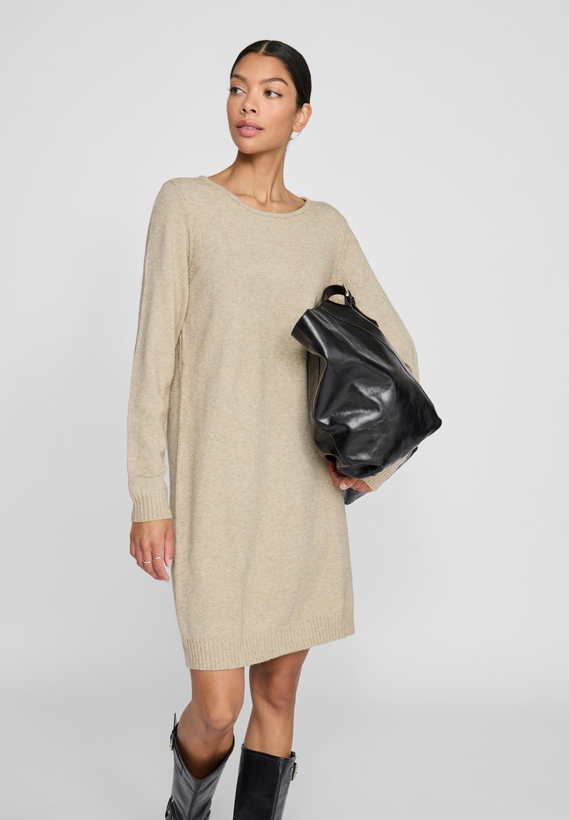 Beige Strickkleid mit langen Ärmeln, rundem Ausschnitt und geripptem Saum. Das Model hält eine schwarze Ledertasche und trägt kniehohe schwarze Stiefel.