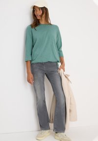 Femme portant un haut à manches longues teal, un jean gris, des baskets beiges et une casquette beige tenant un manteau en peluche couleur crème contre un mur blanc.