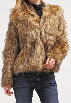 Kobieta w brązowej kurtce z sztucznego futra narzuconej na czarny golf i ciemne dżinsy, stojąca z jedną ręką przy dolnej krawędzi kurtki.