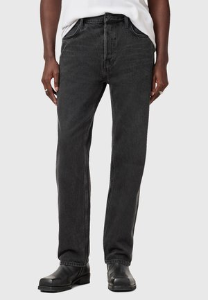 Jeans en denim noir avec une coupe droite, cinq poches et une fermeture à bouton. Texture légèrement délavée avec des coutures contrastantes ; porté avec des chaussures noires.