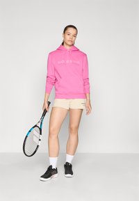 Rosa hettegenser med teksten "BJÖRN BORG", khaki-shortser, svarte treningssko, holder en tennisracket, på en enkel lys bakgrunn.