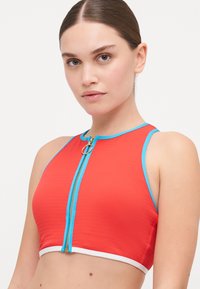 Red crop top with a blue zip detail and white trim. Features a textured fabric and a fitted design with a high neckline and sporty accents.