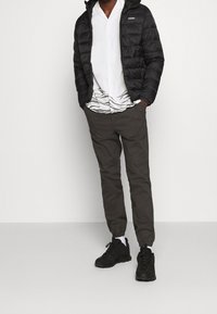 Czarna kurtka puchowa, biała koszula z krótkim rękawem z wzorem fal, oliwkowozielone spodnie dresowe, czarne trampki, stojący na neutralnym tle.