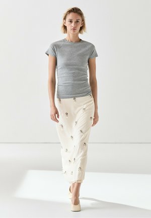 Femme marchant avec un haut gris à manches courtes, une jupe blanche avec de petits motifs floraux et des chaussures couleur crème sur un fond blanc uni.