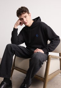 Schwarzer Hoodie mit einem blauen runden Logo auf der Brust. Das Model trägt dunkelgraue Jeans und schwarze Schuhe und sitzt auf einem Holzstuhl.