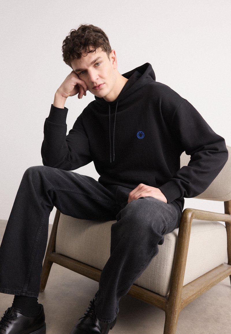 Schwarzer Hoodie mit einem blauen runden Logo auf der Brust. Das Model trägt dunkelgraue Jeans und schwarze Schuhe und sitzt auf einem Holzstuhl.