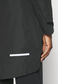 Giacca impermeabile nera con una texture liscia, caratterizzata da una striscia riflettente bianca all'orlo e una regolazione con Velcro sulla manica.