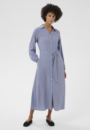 Woman standing wearing a long-sleeve, buttoned, blue patterned midi dress with a waist tie and black slip-on shoes.