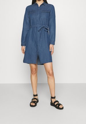 Žena v džínových šatech po kolena s dlouhými rukávy, předními kapsami, páskem uvázaným v pase a černých sandálech s přezkami a kotníkovým páskem.