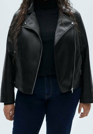 Femme portant une veste en cuir noire avec des fermetures éclair argentées sur un haut noir et un jean bleu foncé, debout devant un fond uni.