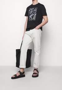 Hombre vistiendo una camiseta gráfica negra, pantalones enrollados color blanco roto, sandalias negras, sosteniendo una bolsa tote negra, de pie contra un fondo blanco.