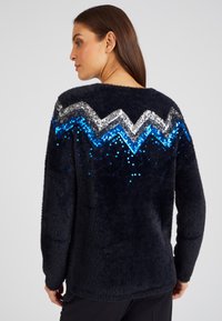 Dunkelblauer, flauschiger Pullover mit einem Zickzackmuster aus silbernen und blauen Pailletten. Lange Ärmel und eine lockere Passform, mit einer weichen Textur.