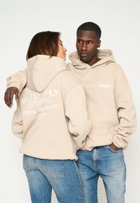 Felpe beige con tasche frontali e cordini. Sul retro presenta un logo e del testo. I modelli indossano jeans azzurri chiari. Tessuto morbido e casual.