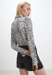Donna con capelli biondi che indossa una blusa a maniche lunghe bianca e nera a pois e pantaloni corti neri a vita alta, posando di lato contro uno sfondo bianco.