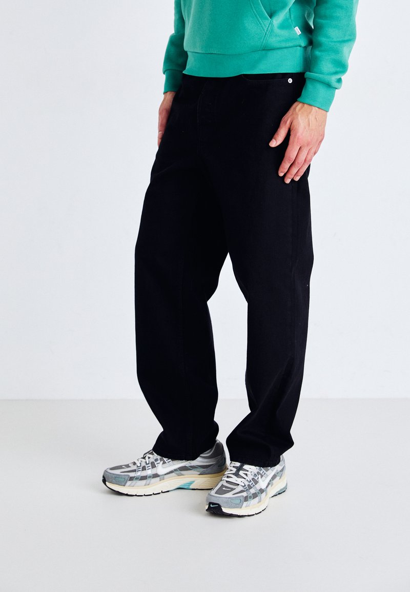 Black denim jeans with a straight leg fit, front pockets, and a subtle logo detail, paired with gray and white running shoes.