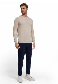 Hellbeige Pullover mit Rundhalsausschnitt, langen Ärmeln und gerippten Bündchen, kombiniert mit marineblauen Anzughosen und weißen Sneakers.